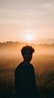 Young Caucasian man standing in a misty golden field at sunrise, soft fog drifting over meadow grass, warm orange sky glowing behind him.