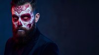 Bearded man with Day of the Dead sugar skull face paint makeup in red and white colors against dark blue background for Halloween costume design.