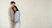 Happy interracial couple embracing tenderly against neutral background, young Caucasian man and Asian woman in casual linen clothing showing love and connection.