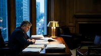 Executive businessman working late at desk in modern office with city skyline view through windows, focused on paperwork and documents during evening hours.