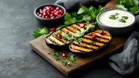 Grilled eggplant slices with charred marks on wooden board, served with yogurt sauce, pomegranate seeds and fresh herbs against dark background.
