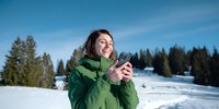 Young woman in green winter jacket smiling while using smartphone in snowy mountain landscape with pine forest background.