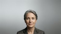 Mature woman with gray hair and neutral expression wearing olive green jacket against gray background, suitable for corporate, business, and professional portraits.