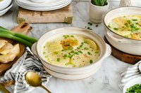 Creamy potato leek soup garnished with chives and olive oil in ceramic bowl on marble surface, surrounded by fresh ingredients and table setting.