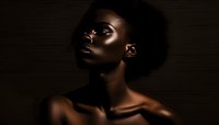 African woman with natural afro hair in dramatic dark lighting against black background, artistic portrait highlighting facial features and glowing skin texture.