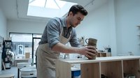 Male carpenter with dark hair and Mediterranean ethnicity working on wooden furniture in bright studio workshop, sanding or finishing wood surface with tools and materials visible.