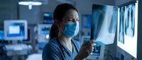Female Asian doctor in medical mask examining chest X-ray films on lightbox in modern hospital radiology department during night shift healthcare work.