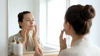 Woman with dark hair in bun applying skincare product to face while looking in mirror. Caucasian female, thirties, performing morning beauty routine in bright bathroom with natural light.