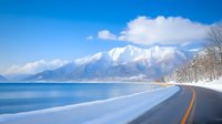 Snow-covered coastal road curves along tranquil blue lake with majestic snow-capped mountains rising in background under bright winter sky and white clouds.