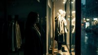 Woman with long dark hair and medium skin tone browsing illuminated storefront window display at night, featuring mannequin wearing beige coat and black pants, urban shopping scene for retail and fashion concepts.