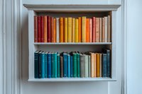 Books arranged by color on white wooden shelves, featuring spectrum from red and yellow tones on top shelf to blue and green volumes below, creating rainbow bookshelf display.