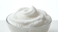 Creamy white moisturizer or cosmetic cream with smooth swirled texture in clear glass bowl on white background for skincare and beauty products.