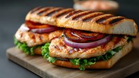 Grilled chicken sandwich with bacon, lettuce and red onion on toasted bun served on wooden board. Fresh fast food meal ready for lunch or dinner service.