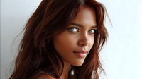Young woman with dark wavy hair and glowing natural makeup gazes confidently at camera against light background, showcasing fresh, radiant complexion and modern beauty look.
