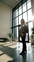 Woman in business casual attire standing in modern office lobby with floor-to-ceiling windows, natural sunlight, and contemporary furniture. Corporate professional environment.