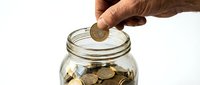 Male hand with medium skin tone dropping a gold coin into a glass jar filled with coins, symbolizing savings, investment, and financial growth.