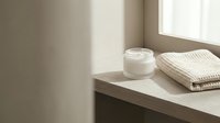 Minimalist bathroom shelf with white cream jar and folded towel near window with natural light, spa and wellness concept.