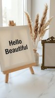 Motivational canvas with Hello text on wooden easel beside pampas grass vase and vintage frame in bright modern interior for home decor and inspiration.