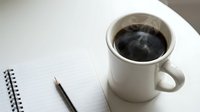 Black coffee in white mug next to open notebook and pen on white desk, overhead view for office, workspace, and productivity concepts.