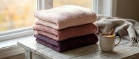 Folded soft towels in neutral and warm tones stacked on wooden table near window with ceramic mug and linen cloth, suitable for home decor and lifestyle content.