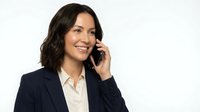 Young woman with dark wavy hair and medium complexion wearing navy blazer and white shirt, speaking on mobile phone against white background.