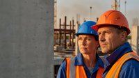 Construction workers in safety helmets and high-visibility vests at building site during golden hour with concrete wall and scaffolding background.