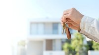 Male hand holding house keys with leather keychain against blurred residential building background, symbolizing homeownership and real estate success.