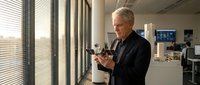 Senior man with gray hair, Caucasian ethnicity, wearing dark business casual attire, holding and examining a small mechanical device or component in a modern office environment near windows.