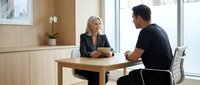Professional blonde businesswoman in suit meeting with male colleague at modern office table discussing work matters in bright contemporary workspace.