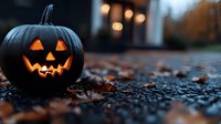Carved jack-o-lantern pumpkin with glowing face sits on autumn pavement surrounded by fallen leaves creating spooky Halloween atmosphere for seasonal decorations.