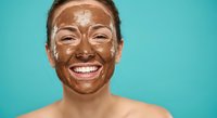 Young African American woman with chocolate face mask smiling joyfully during skincare routine against turquoise background for beauty and wellness content.