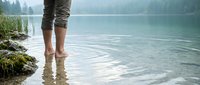 Person standing in shallow mountain lake water with bare feet and rolled up pants enjoying peaceful nature reflection and tranquil forest scenery.