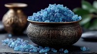 Blue bath salt crystals in ornate metal bowl against dark background with decorative elements and soft natural lighting for spa and wellness concept.