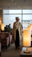 Elegant senior businesswoman walking through modern airport lounge wearing beige trench coat and glasses, carrying leather handbag for luxury travel concepts.
