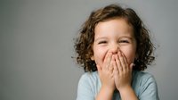 Happy young child with curly hair covering mouth with hands while laughing, expressing joy and playfulness in casual light blue clothing against neutral background.