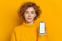 Young woman with curly hair in yellow sweater showing blank smartphone screen against vibrant yellow background for product mockup or app promotion.