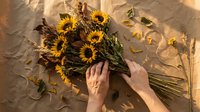 Hands arranging fresh sunflowers on textured beige paper background, overhead flat lay composition for floral design and creative projects.