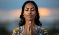 Young Hispanic woman with closed eyes in prayer position wearing floral dress against sunset sky background, expressing peace, spirituality and mindfulness.