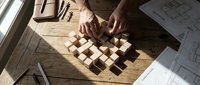 Hands arranging wooden building blocks on rustic table with architectural blueprints and natural sunlight streaming through window for creative design process.