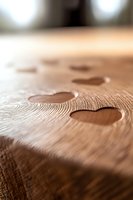 Water drops in heart shape on wooden surface with natural grain texture, macro photography showing detailed wood structure in warm brown tones.