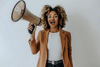 Young African American woman with curly hair wearing brown blazer and white top holds megaphone while expressing excitement against light background.