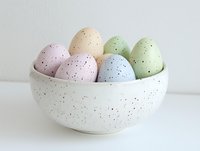 Speckled pastel Easter eggs in white ceramic bowl against light background, featuring soft pink, blue, peach and mint green colors with delicate spotted pattern.