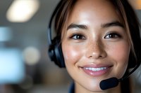 Young Asian female customer service representative wearing headset smiles warmly while working in modern office environment. Professional portrait with soft bokeh background.