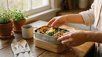 Caucasian woman in casual beige shirt holding lunch container with grilled vegetables and grains at wooden table near window, healthy meal prep and eating concept.