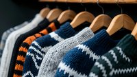 Cozy knitted winter sweaters with traditional Nordic patterns hanging on wooden hangers in closet, featuring navy blue, orange, and grey color combinations.