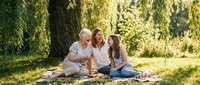 Happy multi-generational family enjoying picnic together in sunny park. Grandmother, mother and teenage daughter bonding outdoors on blanket under willow tree.