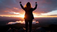 Young woman with raised arms celebrating success on mountain peak at dramatic sunset, silhouetted against orange sky and mountain landscape with clouds below.