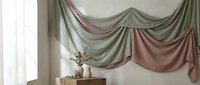 Elegant draped fabric swags in muted sage green and dusty rose tones hang from window, creating sophisticated layered curtain design with dried botanical arrangement on wooden table below.