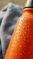 Close-up of condensation droplets on an orange beverage bottle with blurred towel background, suitable for refreshment and hydration advertising.