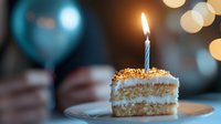 Slice of layered cake with lit birthday candle on plate against blurred festive background with bokeh lights.
